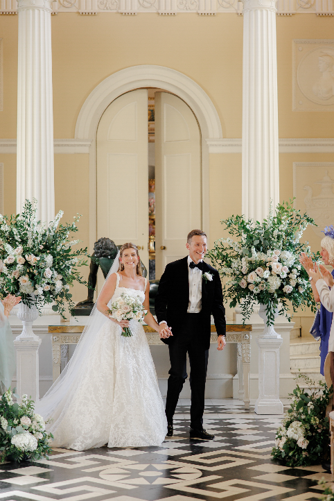 Newly married Olivia and Jack walk between large white and blush floral pedestal arrangements inside Syon House