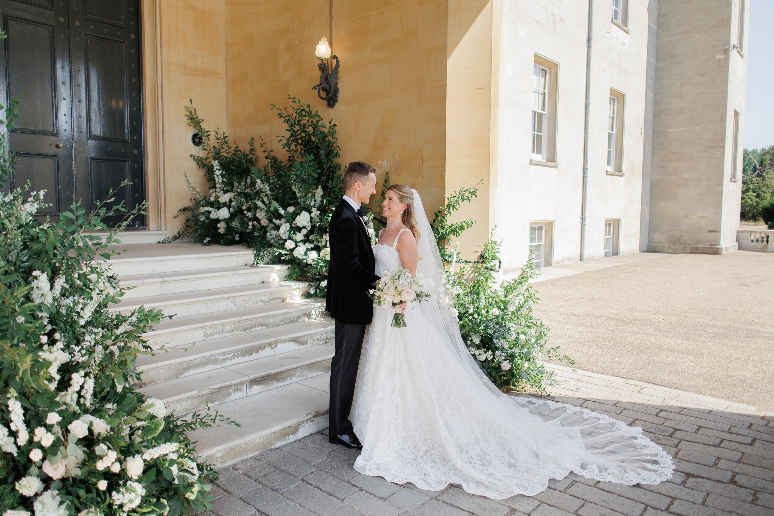 Olivia and Jack beside abundant white and blush floral installations on the steps of Syon House